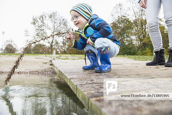 Kleiner Junge hockt auf einem Landungssteg am See und hält einen Zweig ins Wasser  mit seiner Mutter im Hintergrund