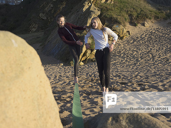 Teenage girl balancing along a slackline on a beach assisted by her sister