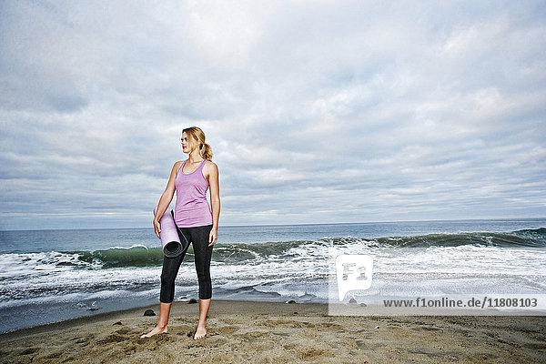 Caucasian woman standing on ocean beach holding exercise mat