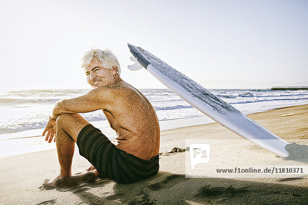 Älterer kaukasischer Mann am Strand sitzend mit Surfbrett