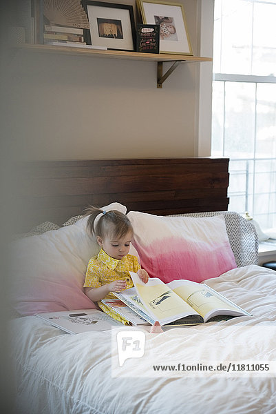 Caucasian baby girl sitting on bed reading book