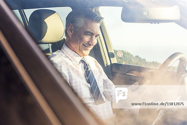 Lächelnder kaukasischer Geschäftsmann sitzt im Auto und schreibt eine SMS auf seinem Handy
