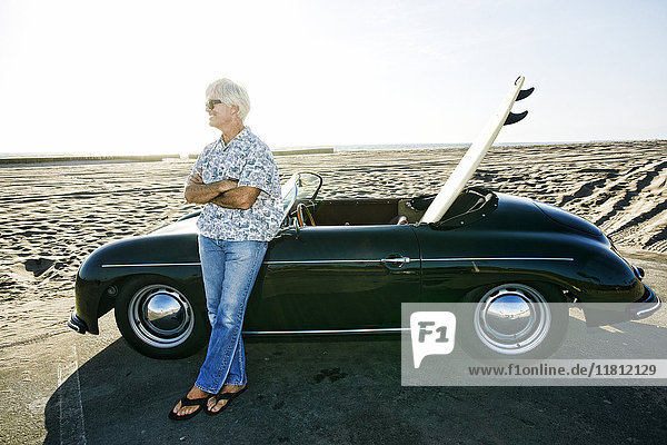 Älterer kaukasischer Mann lehnt an Cabrio mit Surfbrett am Strand