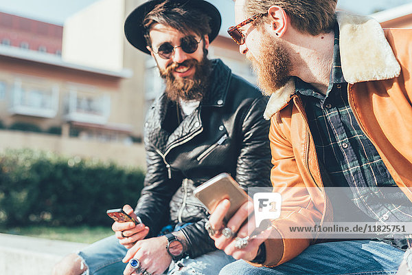 Zwei junge männliche Hipster sitzen mit Smartphones an der Wand