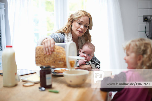 Mutter hält einen kleinen Jungen  sitzt mit der kleinen Tochter am Küchentisch und schüttet Müsli in eine Schüssel für die Tochter