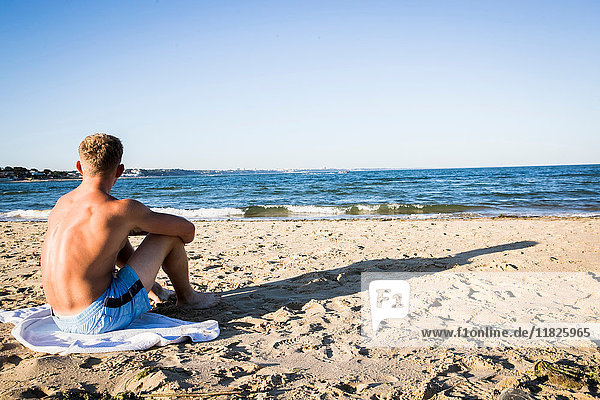Junger Mann sitzt am Strand und schaut aufs Meer