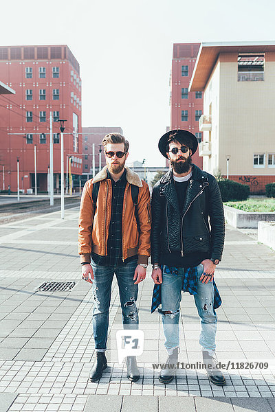 Portrait of two young male hipster friends standing in city housing estate  full length