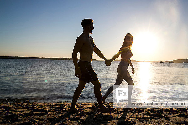 Hinterleuchteter junger Mann und Freundin spazieren Hand in Hand am Strand bei Sonnenuntergang