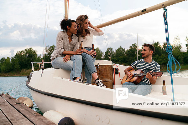 Three friends relaxing on moored sailing boat  drinking beer  man playing guitar
