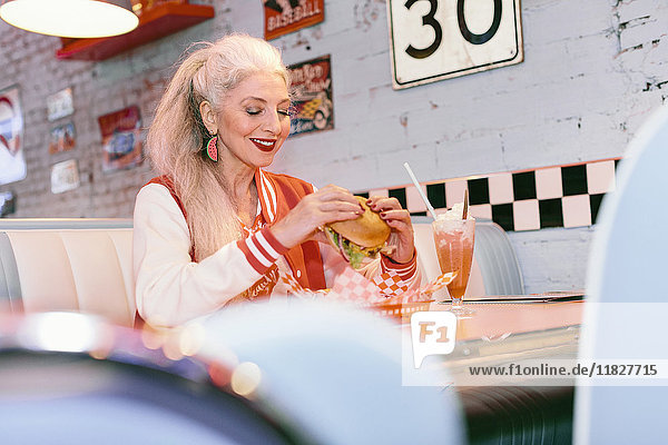 Reife Frau in Baseballjacke isst Burger im Diner der 1950er Jahre