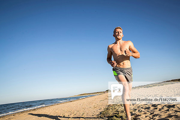 Nacktbrust junger männlicher Läufer läuft am Strand gegen blauen Himmel