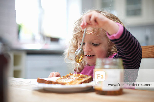 Junges Mädchen sitzt am Küchentisch und träufelt Honig auf Toast