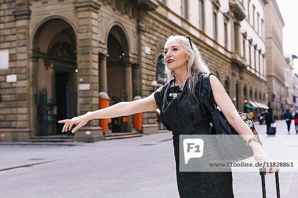 Mature woman with long grey hair with wheeled suitcase hailing a cab in Florence  Italy