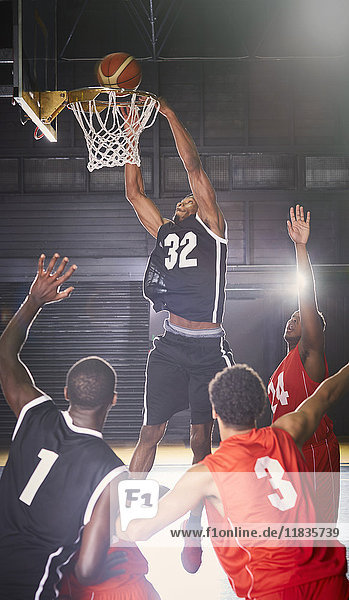 Junger Basketballspieler taucht den Ball in einen Reifen mit Verteidigern darunter.