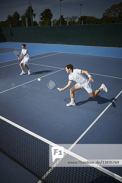 Junge Tennisspieler,  die auf dem sonnigen,  blauen Tennisplatz nach dem Ball greifen.