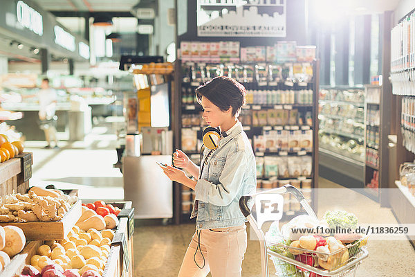 Young woman with headphones using cell phone  grocery shopping in market