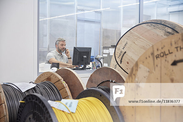 Male supervisor working at computer and talking on ell phone behind spools in fiber optics factory