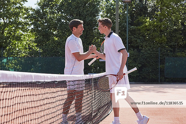 Young male tennis players shaking hands in sportsmanship over net on sunny clay tennis court