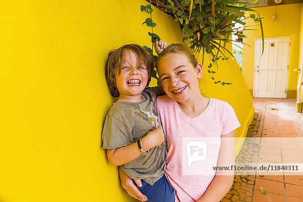 Kaukasisches Mädchen lehnt an einer gelben Wand und hält ihren Bruder