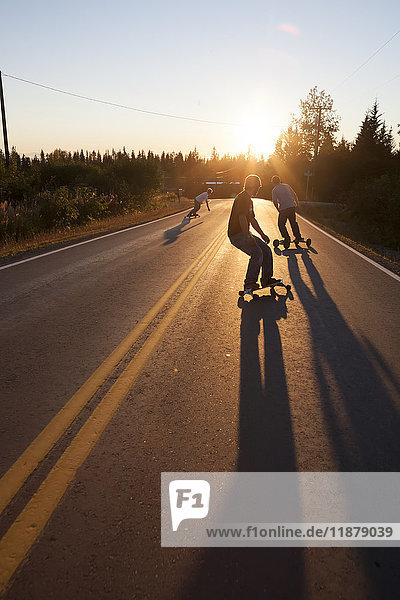 'Three young men skateboarding down a road at sunset; Homer  Alaska  United States of America'