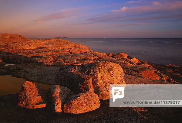 Das Licht des Sonnenuntergangs taucht eine felsige Küste und einen entfernten Leuchtturm ein; Sambro Island  Nova Scotia  Kanada'.