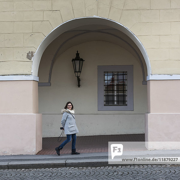 'A woman strolls by a building under an archway with a smile  holding a camera behind her back; Prague  Czech Republic'