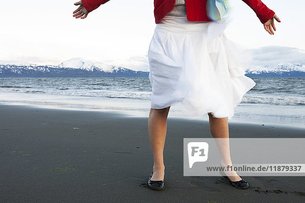 Eine junge Frau steht mit ausgestreckten Armen am Strand und trägt einen weißen Rock und eine rote Jacke; Homer  Alaska  Vereinigte Staaten von Amerika'.
