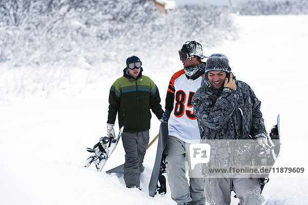'Three snowboarders walking up a trail in deep snow with their snowboards  one snowboarder talking on a cell phone; Alaska  United States of America'
