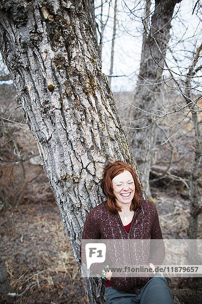 'Portrait of a woman with red hair leaning on a tree and laughing with eyes closed; Homer  Alaska  United States of America'