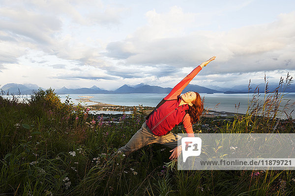 Eine Frau macht Yoga auf einer Wiese mit Blick auf Homer Spit  Kachemak Bay und die Kenai Mountains im Hintergrund; Homer  Alaska  Vereinigte Staaten von Amerika'.