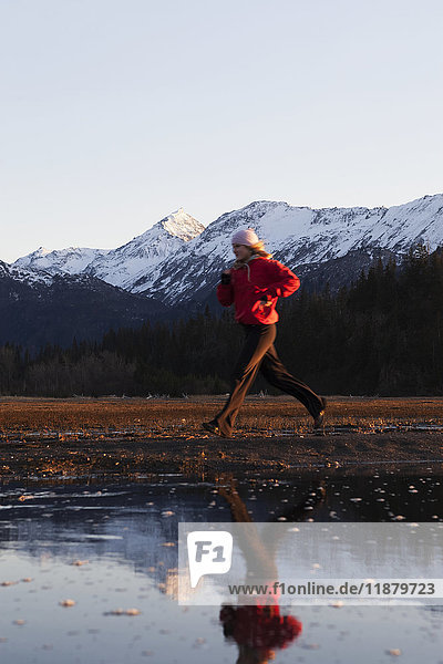 Eine junge Frau läuft am Wasser entlang  mit ihrem Spiegelbild im Wasser und den Kenai Mountains im Hintergrund  Kachemak Bay State Park; Alaska  Vereinigte Staaten von Amerika'.