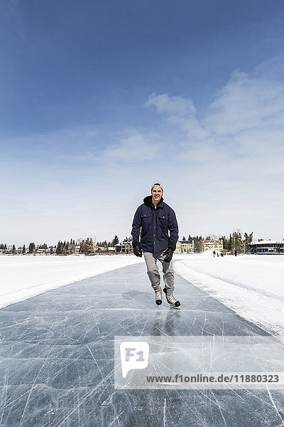 Mann beim Schlittschuhlaufen auf frisch präpariertem Eis auf einem See mit Häusern im Hintergrund und blauem Himmel; Calgary  Alberta  Kanada'.