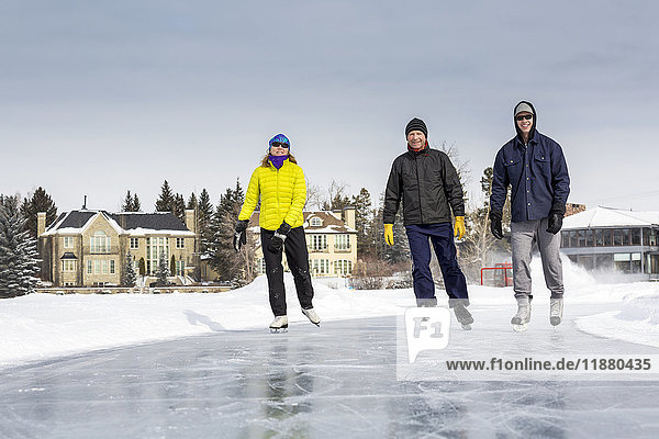 'Two males and one female skating on freshly groomed ice on pond with houses in the background; Calgary  Alberta  Canada'