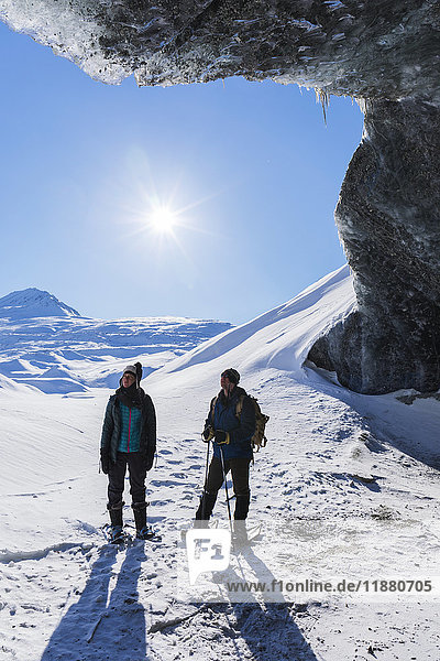'A couple snowshoeing and standing beneath the entrance to an ice cave on a sunny winter day at Canwell Glacier in the Alaska Range; Alaska  United States of America'