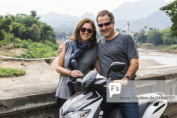 'A couple stand posing by a motorcycle with a mountainous landscape in the distance; Luang Prabang  Luang Prabang Province  Laos'