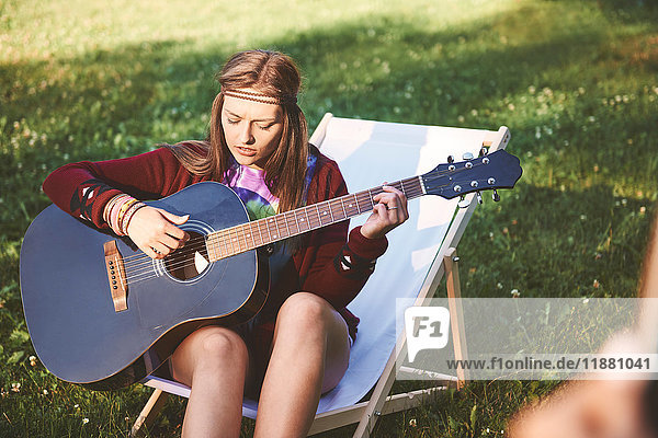 Junge Boho-Frau sitzt auf Liegestuhl und spielt Akustikgitarre beim Festival