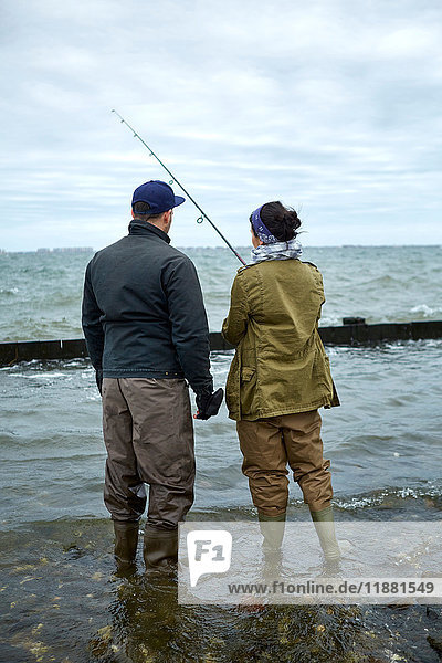 Rückansicht eines jungen Mannes  der knöcheltief im Wasser steht und seiner Freundin das Meeresangeln beibringt