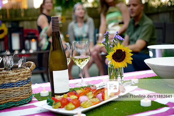 Group of people at garden party  bottle of wine and vegetable kebabs in foreground