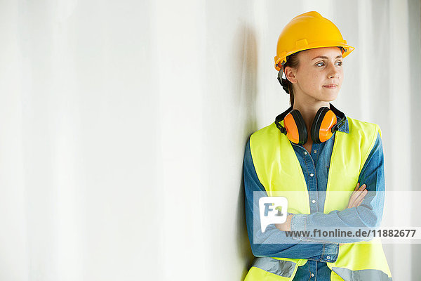 Junge Frau  trägt Hi-Vis-Weste und Schutzhelm  lehnt an der Wand  Arme verschränkt