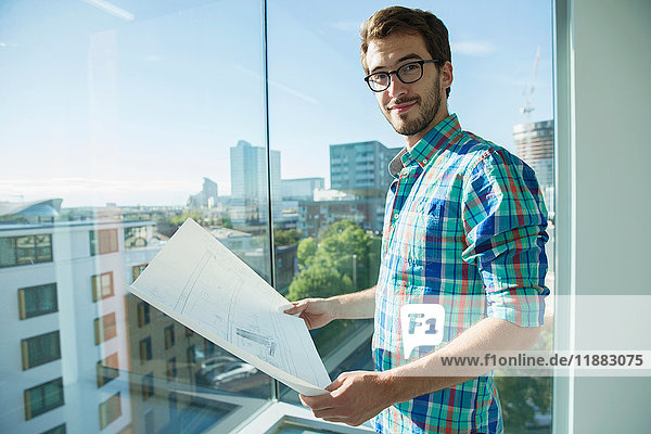 Porträt eines Mannes der neben einem Bürofenster steht und auf Pläne schaut