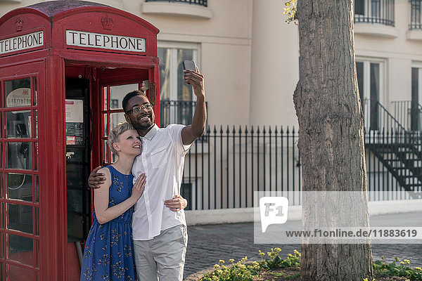 Junges Paar steht neben der roten Telefonzelle  schaut auf und lächelt