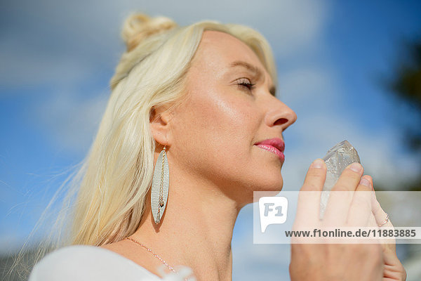 Mittelgroße erwachsene Frau meditiert und hält einen Kristall gegen den blauen Himmel