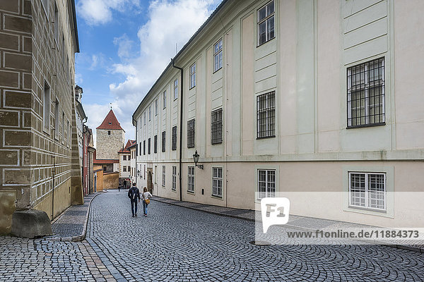 'A couple walk down a street with patterned paving stones between buildings; Prague  Czech Republic'