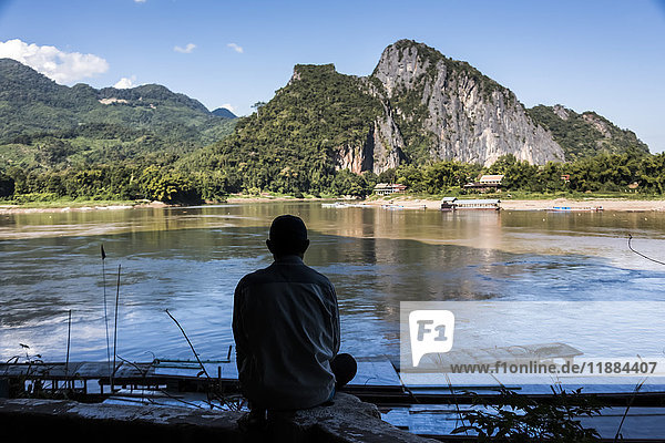 'A man sits on the edge of the river looking out at the mountainous landscape and tranquil river; Pak Ou  Luang Prabang Province  Laos'