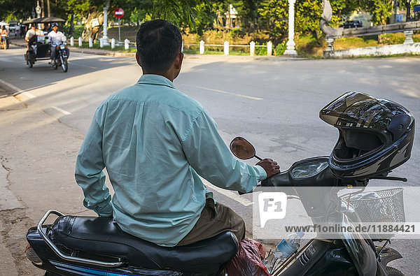'A man sits on his motorcycle at the side of the road as a cycle rickshaw approaches; Krong Siem Reap  Siem Reap Province  Cambodia'
