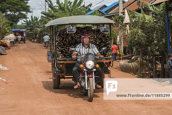 'A man rides a motorcycle pulling a cart full of cut wood; Siem Reap Province Cambodia'