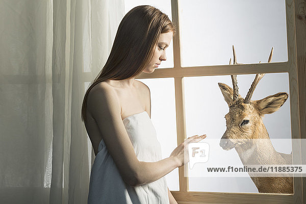 Nachdenkliche Frau in eine Decke gehüllt  die Hirsche ansieht  während sie am Fenster steht.