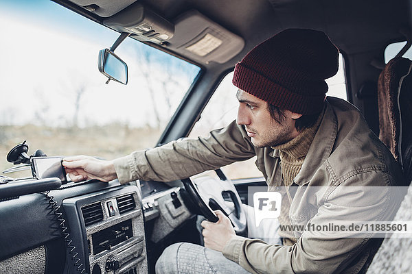 Man wearing knit hat and adjusting smart phone on dashboard of sports utility vehicle
