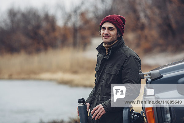 Lächelnder Mann hält isolierten Getränkebehälter  während er am Seeufer neben einem Geländewagen steht