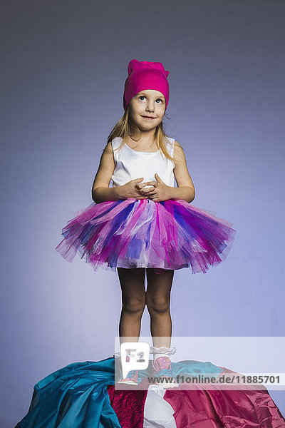 Cute girl wearing tulle skirt and knit hat standing on fabrics heap against purple background